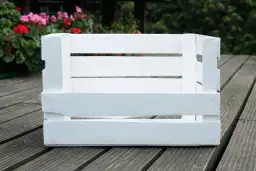 Biała drewniana skrzynka, idealna baza, by dowiedzieć się, jak zrobić legowisko dla kota. W tle kwitnące kwiaty.