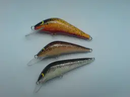 Trzy spinningowe przynęty na pstrąga w różnych kolorach: żółto-pomarańczowa, brązowa i czarno-biała.