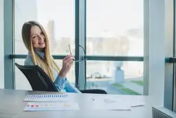 Uśmiechnięta blondynka w biurze, trzymająca okulary, zastanawia się, kiedy można wziąć urlop bezpłatny.