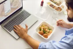 Mężczyzna je dietetyczny lunch z pudełka, pracując na laptopie.