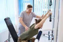 Fizjoterapeuta pomaga mężczyźnie w ćwiczeniach rehabilitacyjnych. Dostępne są bezpłatne wyjazdy dla osób niepełnosprawnych, by mogły skorzystać z takich terapii.