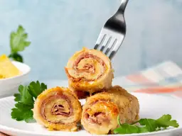 Roladki drobiowe z serem, zawinięte z szynką, prezentują się apetycznie na białym talerzu. Jedna z nich jest nabita na widelec.