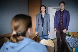 Dwoje ludzi stoi w pokoju, patrząc na trzecią osobę zza kadru.