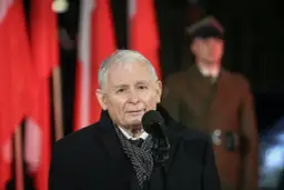 Jarosław Kaczyński przemawia przy mikrofonie na tle polskich flag i żołnierza.