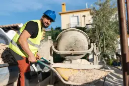 Budowlaniec w kamizelce i kasku obsługuje betoniarkę. Zastanawiasz się, ile waży betoniarka?