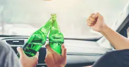 Dwie osoby w samochodzie wznoszą toast zielonymi butelkami piwa. Zastanawiają się, ile można wypić alkomat.