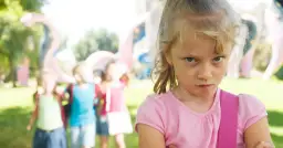 Zagniewana dziewczynka, bunt 7 latka, na tle bawiących się dzieci i placu zabaw.
