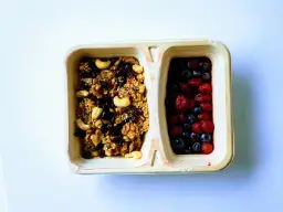 Dwa przedziały pojemnika: jeden z granolą i orzechami, drugi z jagodami i truskawkami.