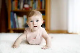 Niemowlę na brzuchu na białym dywanie, z niebieskimi oczami, na tle regału z książkami.