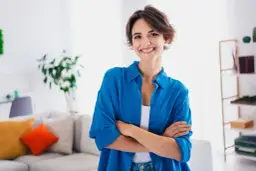 Uśmiechnięta kobieta w niebieskiej koszuli stoi ze skrzyżowanymi rękami w jasnym wnętrzu.