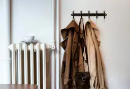Biały kaloryfer z filiżanką kawy, obok płaszcze na wieszaku. Pomysł, jak ukryć rury od kaloryfera, może być inspirowany tym minimalistycznym wnętrzem.