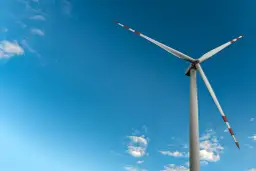 Mała elektrownia wiatrowa z białymi łopatami i czerwono-białymi końcówkami na tle błękitnego nieba z chmurami.