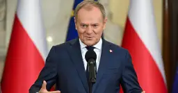 Donald Tusk w garniturze przemawia przy mikrofonie na tle flag Polski i UE.