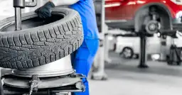 Mechanik w niebieskim kombinezonie trzyma oponę na tle warsztatu samochodowego.