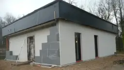 Budowa domu ze styropianu w toku. Ściany z bloczków, częściowo otynkowane, dach przykryty czarną folią.