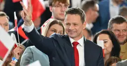 Rafał Trzaskowski w garniturze i czerwonym krawacie, pozdrawia tłum, unosząc rękę ze znakiem zwycięstwa.