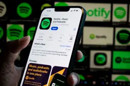 Dłoń trzyma smartfon z otwartą aplikacją Spotify. W tle widać ikony Spotify.