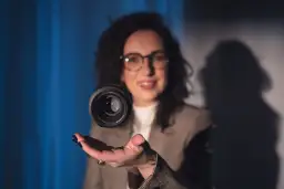 Uśmiechnięta kobieta w okularach unosi obiektyw aparatu nad dłonią. Czy fotograf przechowuje zdjęcia w obiektywie?