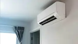Biała jednostka klimatyzacyjna zamontowana na ścianie, obok okna z zasłoną.