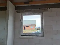 Ściana z bloczków z widocznym oknem i cofniętym nadprożem do rolet. W tle dom w budowie.