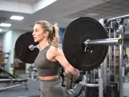 Kobieta z blond kucykiem ćwiczy z ciężarem na sztandze w siłowni.