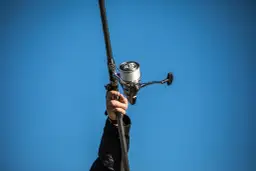 Dłoń trzyma wędkę z nawiniętą plecionką na kołowrotek, na tle błękitnego nieba.