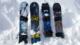 Cztery deski snowboardowe z różnymi grafikami i wiązaniami, leżące na śniegu.