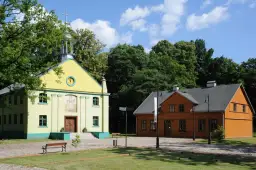 Żółty kościółek i pomarańczowy dom drewniany na tle zieleni i błękitnego nieba.