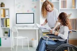 Kobieta na wózku inwalidzkim omawia wykresy z koleżanką. W miejscu pracy, gdzie stopnie niepełnosprawności nie ograniczają, a wręcz dają pewne przywileje, obie panie współpracują.