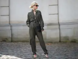 Kobieta w oliwkowym kombinezonie, czarnym pasku i białych sandałach na obcasie, trzymająca czarną kopertówkę.