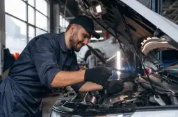 Uśmiechnięty mechanik samochodowy w granatowym kombinezonie sprawdza silnik. Zastanawiasz się, ile zarabia mechanik samochodowy?