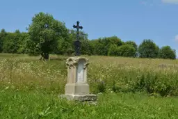 Kamienny postument z krzyżem na wzgórzu porośniętym dzikimi kwiatami. W tle las i błękitne niebo. Dźwiniacz górny.