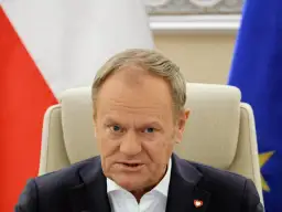 Donald Tusk, z flagą Polski i UE za plecami, w białej koszuli i granatowej marynarce. Czy Donald Tusk ma pochodzenie niemieckie?