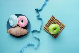 Dwa pączki i rogalik na talerzu obok zielonego jabłka i miarki krawieckiej. Wybór między słodyczami a zdrowiem, między kj a kcal.