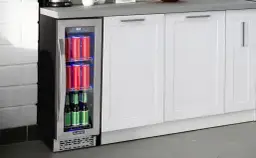 Szklana lodówka na napoje z czerwonymi puszkami i zielonymi butelkami, obok białych szafek kuchennych.