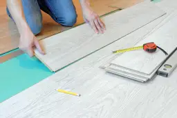 Ręce układają jasne panele podłogowe, obok miarka i poziomica. Widać, jak dobijać panele, by stworzyć idealną podłogę.