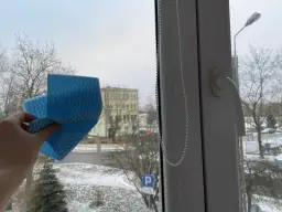 Ręka trzyma niebieską ściereczkę, gotową do mycia okna. Za szybą zimowy krajobraz z drzewami i budynkiem. Czy można myć okna na mrozie?