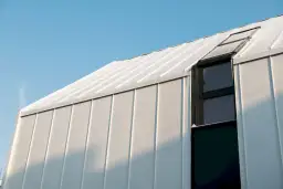 Nowoczesny budynek z białej blachy na rabek, pokryty śniegiem, z ciemnym oknem i świetlikiem.