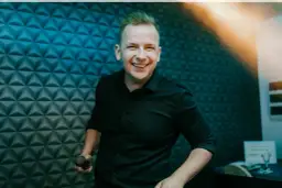 Uśmiechnięty DJ w czarnej koszuli trzyma mikrofon na tle niebieskiej, geometrycznej ściany.