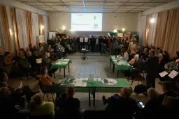 Ludzie siedzą i stoją na spotkaniu, na stole leżą dokumenty i laptop.
