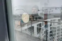 Mężczyzna w czapce wykonuje polerowanie szkła za pomocą maszyny. W odbiciu widać budynki i dźwigi.