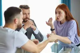 Trzy osoby przy stole, mężczyzna w garniturze i kobieta z rudymi włosami dyskutują, mężczyzna z przodu w białej koszulce.