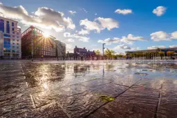 Zachód słońca nad miejską fontanną, odbijający się w mokrej nawierzchni. Czułość filmu uchwycona w złotych promieniach i tańczących kroplach wody.