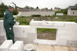 Budowlaniec montuje gotowe nadproża, tworząc otwór okienny. W tle widać ogród i domy.