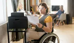 Kobieta na wózku inwalidzkim drukuje dokumenty. Zastanawia się, kto płaci asystentowi osoby niepełnosprawnej, gdy ona sama pracuje.
