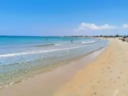 A wide, sandy beach with clear turquoise water and a few people swimming and sunbathing under a bright blue sky.