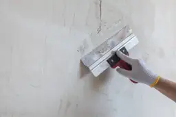Dłoń w rękawicy budowlanej używa kielni do nakładania masy na nierówną, jasną ścianę.