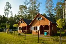 Dwa drewniane domki rekreacji indywidualnej, idealne na wynajem, stoją na zielonej trawie wśród drzew.