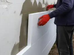 Pracownik w czerwonych rękawicach przykłada styropian do ściany. Trwa gruntowanie elewacji po ociepleniu.