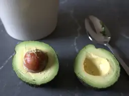 Dwa połówki awokado, jedna z pestką, druga pusta, obok łyżka. Prosty sposób, jak obierać awokado.